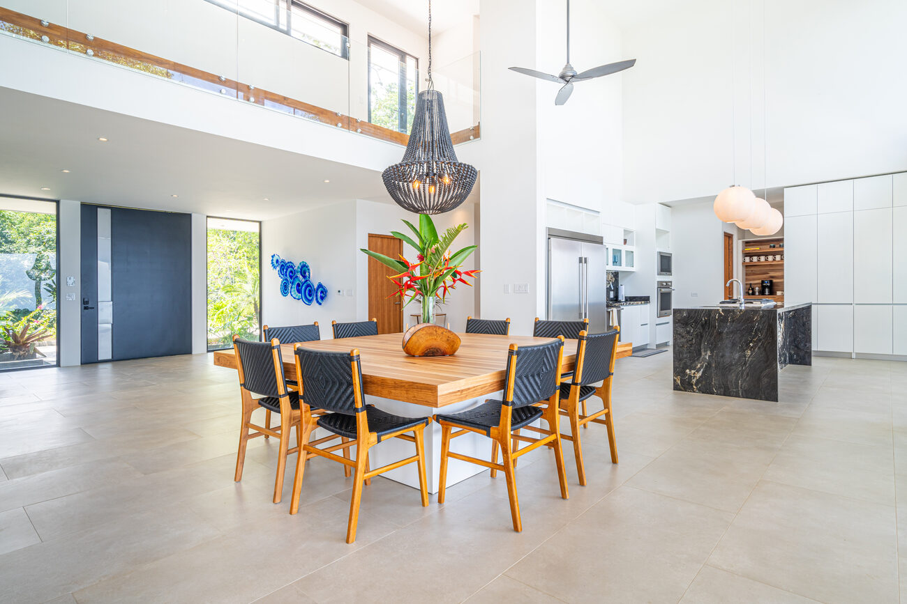 The open dining and kitchen — square teak table, marble island, double-height volume
